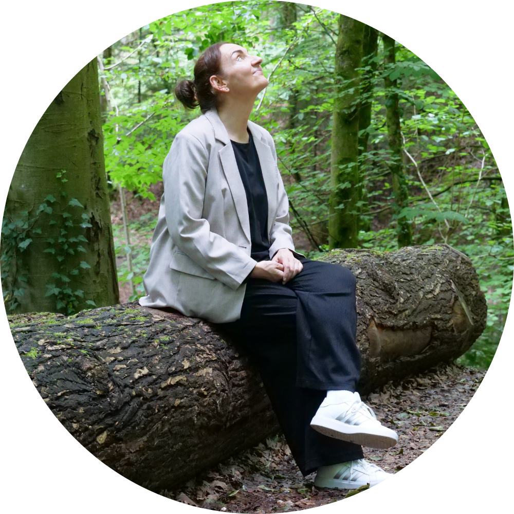 Frau sitzt im Wald auf einem Baumstamm und schaut in die Luft